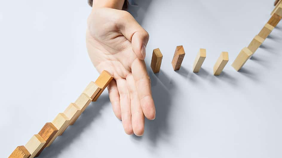 Dettaglio di una mano che blocca il percorso di caduta dei pezzi di un domino di legno.