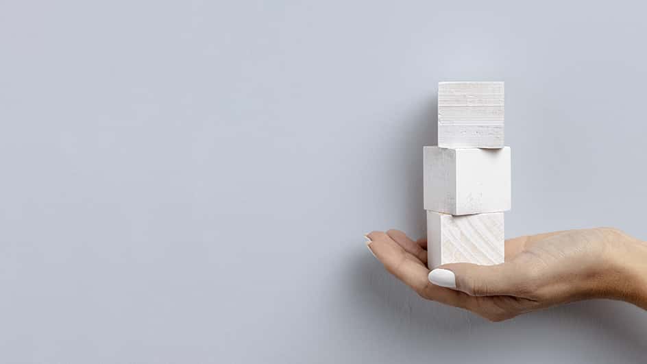 Dettaglio di una mano femminile con smalto bianco che sorregge tre blocchi di legno bianco.